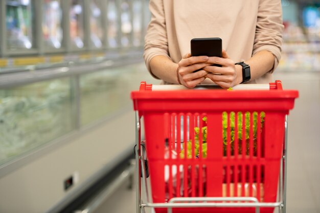 Foto unerkennbare junge frau, die im gang mit einkaufswagen im supermarkt steht und die einkaufsliste auf dem smartphone überprüft