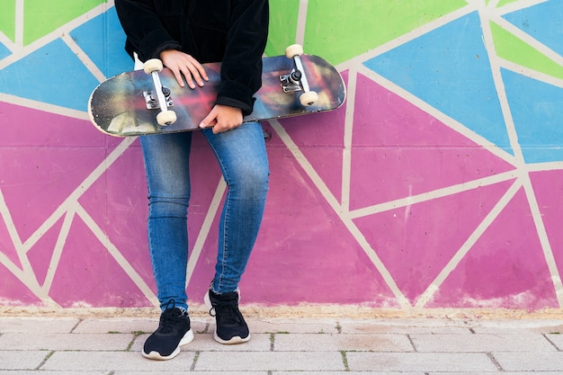 Unerkennbare junge Frau mit Skateboard an eine bunte Wand gelehnt