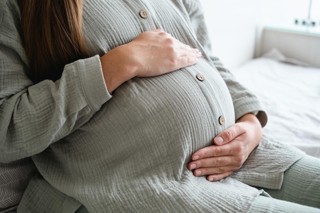 Unerkennbare langhaarige Frau, die zu Hause auf dem Bett liegt und die Hände auf den Bauch legt, glückliche, gesunde Schwangerschaft