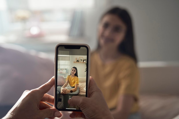 Unerkennbare Person macht mit dem Handy ein Foto von einem weißen Teenager, das auf dem Bett sitzt
