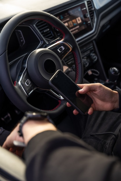 Unerkennbarer Mann, der mit einem Mobiltelefon auf dem Pilotensitz sitzt, dunkle, moderne Kleidung trägt, das Telefon in einer Hand und eine Smartwatch in der anderen hält.