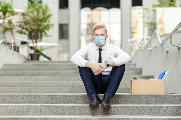 Unfairer Arbeiter mit chirurgischer medizinischer Maske, der auf der Treppe sitzt und ernsthaft in die Kamera schaut