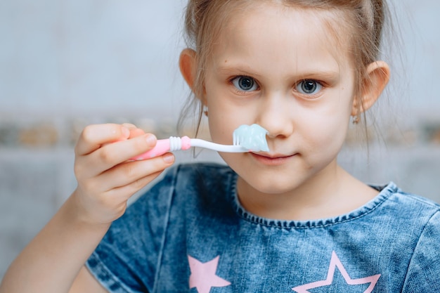 Ungefähres Gesicht eines kleinen Vorschulmädchens mit einer Zahnbürste, die mit Paste in der Hand bedeckt ist Verschwommener Kachelhintergrund