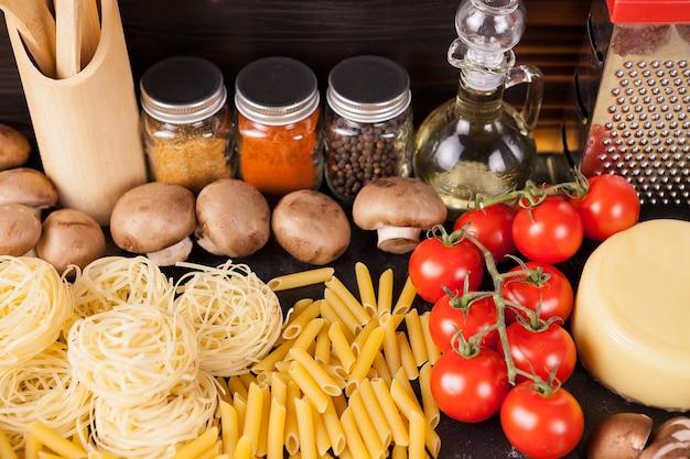 Ungekochte rohe Makkaroni, Nudeln und Spaghetti neben frischem und gesundem Gemüse, verschiedenen Gewürzen und Sonnenblumenöl auf dunklem rustikalem Holzhintergrund