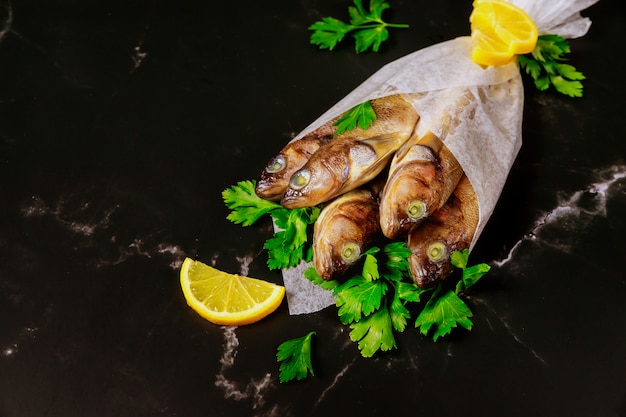 Ungekochter ganzer Fisch mit Petersilie und Zitrone, eingewickelt in Pergamentpapier auf schwarzem Tisch.