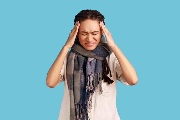 Ungesunde Frau mit schwarzen Dreadlocks, die in einen Schal gewickelt sind, der kalte Schläfen fängt, um Kopfschmerzen zu lindern, hält die Augen geschlossen und hat eine hohe Temperatur Studioaufnahme im Innenbereich isoliert auf blauem Hintergrund