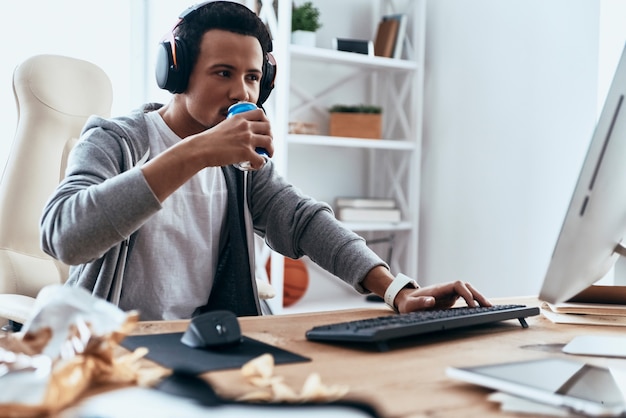 Ungesunder Snack. Konzentrierter junger Mann in Freizeitkleidung mit Computer, während er Zeit zu Hause verbringt