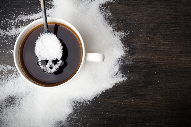 Ungesundes Weißzuckerkonzept. Schädellöffel mit Zucker und Tasse schwarzen Kaffees auf hölzernem Hintergrund