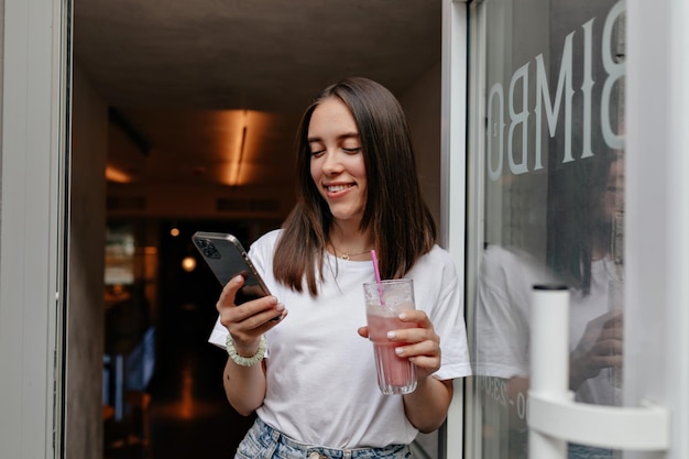 Unglaubliche stilvolle Frau mit kurzer dunkler Frisur hält Smartphone und lächelt, während sie in der Cafeteria steht und Sommersmoothie trinkt