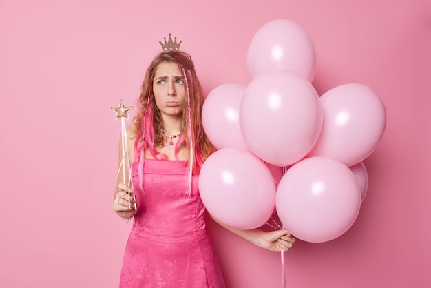 Unglückliche langhaarige junge Frau runzelt die Stirn, hat schlechte Laune und hält einen Zauberstab, einen Haufen aufgeblasener Luftballons, trägt Krone und Kleid isoliert über rosa Hintergrund. Menschen Urlaub Feierkonzept.