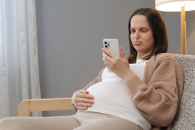 Unglückliche schwangere Frau, die zu Hause ein Smartphone benutzt, die Schwangerschafts-App durchsucht, während sie auf der Couch im Wohnzimmer sitzt, lächelnd, werdende Dame mit unzufriedenem Gesicht, die auf den Telefonbildschirm schaut