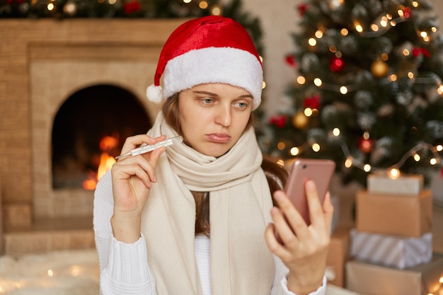 Unglückliche und ungesunde Frau, die im dekorierten Wohnzimmertelefon in den Händen aufwirft, über Smartphone spricht und Thermometer zur Gerätekamera zeigt, enttäuscht zu sein, während der Weihnachtsferien krank zu sein.