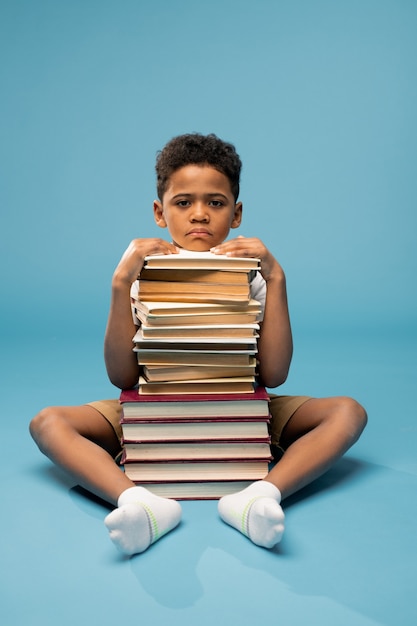 Unglücklicher afrikanischer Junge im Grundschulalter, der mit einem hohen Stapel Bücher vorne auf dem Boden sitzt und sein Kinn darüber hält
