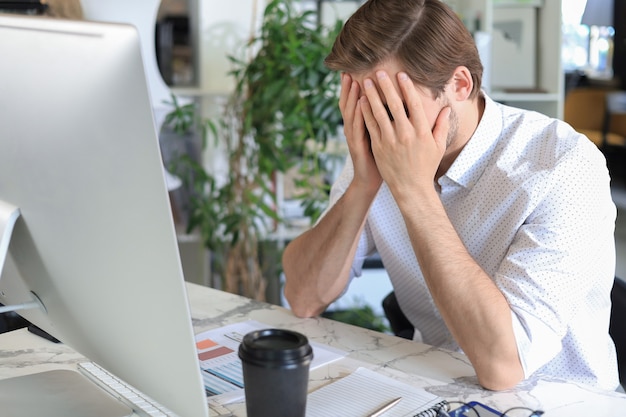 Unglücklicher frustrierter junger Mann, der den Kopf mit den Händen hält und mit dem Computer hinter dem Schreibtisch im Büro sitzt.