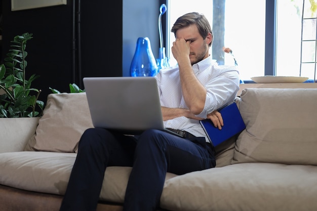 Unglücklicher frustrierter junger Mann, der den Kopf mit der Hand hält und mit Laptop auf dem Sofa sitzt.