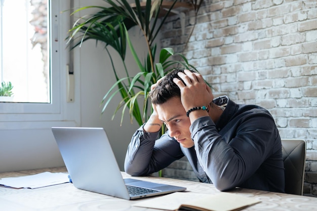 Unglücklicher junger kaukasischer männlicher Arbeiter in Brille schaut auf den Laptopbildschirm, schockiert von dem Gadget