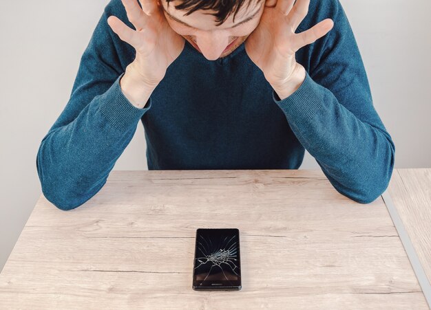 Unglücklicher junger Mann, der sein Smartphone mit einem kaputten Bildschirm betrachtet