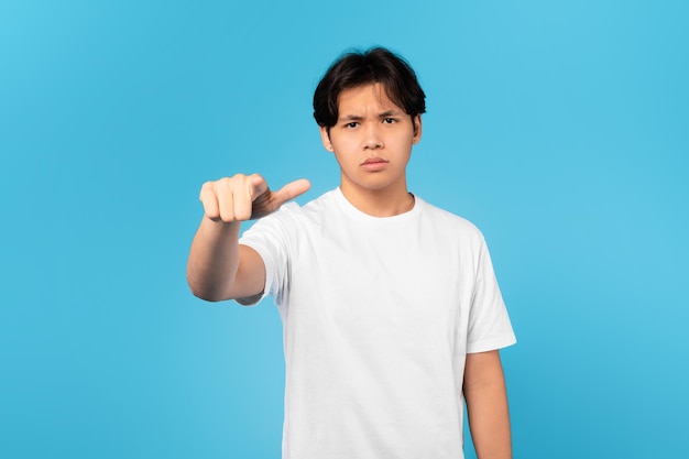 Unglücklicher koreanischer Teenager, der mit dem Finger die Stirn runzelt und über blauem Hintergrund zeigt