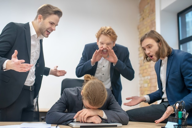 Unglücklicher Mann am Tisch mit gesenktem Kopf vor dem Laptop und drei schreienden wütenden Kollegen im Büro