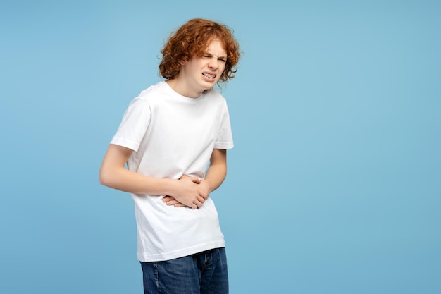 Unglücklicher rothaariger Junge mit Magenschmerzen isoliert auf blauem Hintergrund