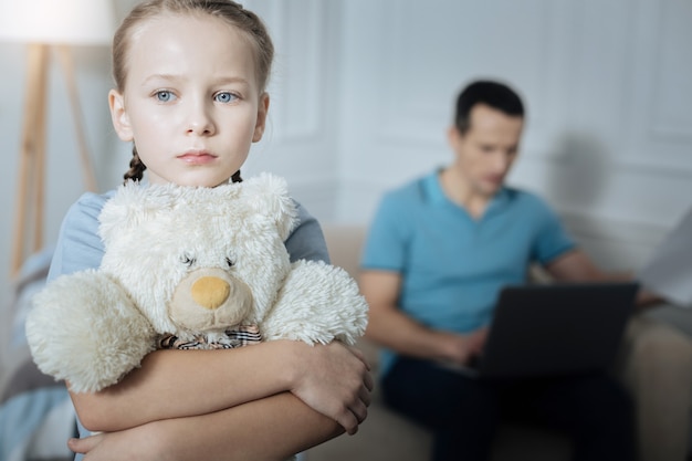 Unglückliches blauäugiges blondes kleines Mädchen, das ihr Spielzeug hält und im Raum steht, während ihr Vater im Hintergrund arbeitet