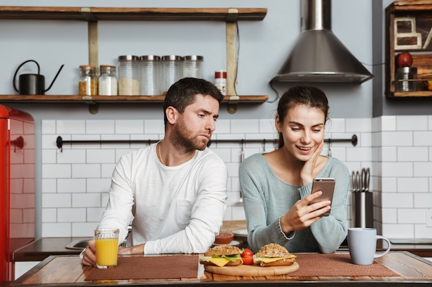 Unglückliches junges Paar, das während des Mittagessens zu Hause in der Küche sitzt, ein Problem hat und Handy hält