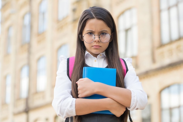 Unglückliches kleines Kind mit Brille hält Lernbuch auf dem Schulhof im Freien zurück zur Schule