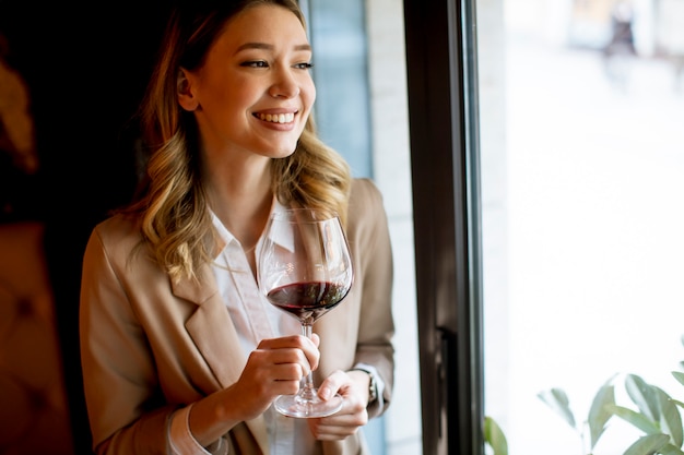 Única jovem mulher com um copo de vinho tinto em pé perto da janela e olhando de lado