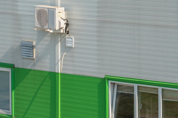 Unidad de aire acondicionado exterior en la pared de una fábrica o fábrica La pared exterior de la empresa está hecha de perfiles de metal blanco y verde con tuberías de drenaje y grandes ventanales para copiar texto