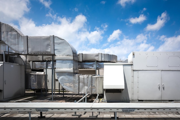 Unidade de ar condicionado quadrada no telhado com um ventilador redondo Ao fundo recuando gradualmente outras unidades que estão fora de foco No lado direito céu azul claro e espaço comercial