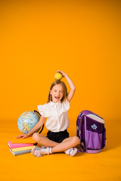 El uniforme de colegiala se sienta con útiles escolares y un globo y sostiene una manzana en la cabeza sobre un fondo amarillo con una copia del espacio