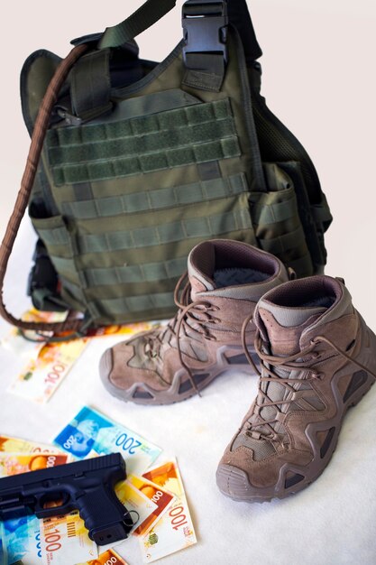 Uniforme y equipo militar. Chalecos antibalas, pistola negra y botas con billetes de Israel New Shekels vista superior. Montón de billetes de dinero israelíes y pistola sobre fondo blanco. Concepto visual de soldado