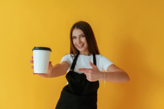 Uniforme de trabajador de café blanco y negro sosteniendo una taza de bebida Mujer joven contra un fondo amarillo