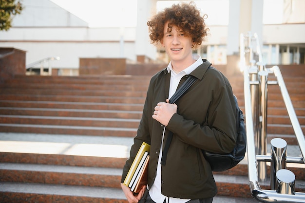 UniversidadSonriente joven estudiante hombre sosteniendo un libro y una bolsa sobre un fondo universitario Joven estudiante sonriente al aire libre Estilo de vidaCiudadEstudiante