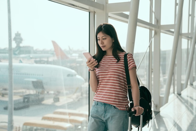 universitária chinesa no aeroporto verificando a agenda no smartphone enquanto caminhava com bagagem de mão no aeroporto de hong kong indo para o portão de embarque. mulher usando o celular para conversar nas férias de verão