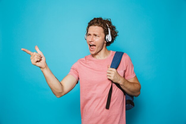Universitário com cabelo encaracolado, usando mochila, ouvindo música através de fones de ouvido e apontando o dedo para o lado
