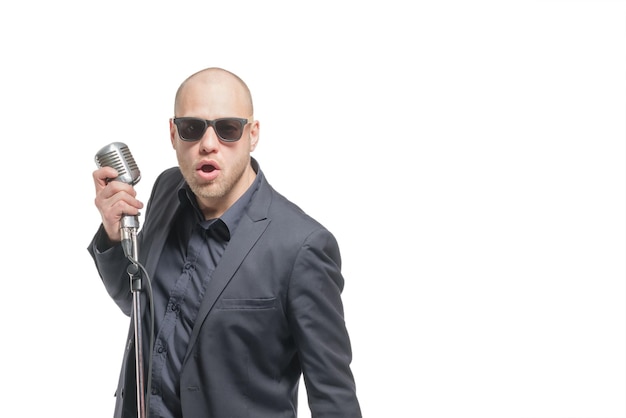 Unrasierter Glatzkopf in grauem Anzug und Sonnenbrille, der ein Mikrofon hält und isoliert singt