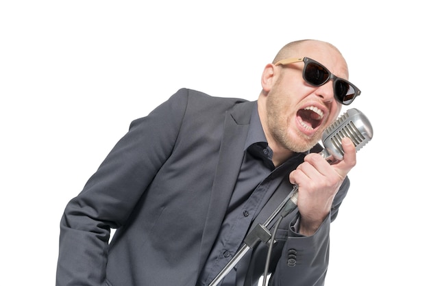 Unrasierter Glatzkopf in grauem Anzug und Sonnenbrille, der ein Mikrofon hält und isoliert singt