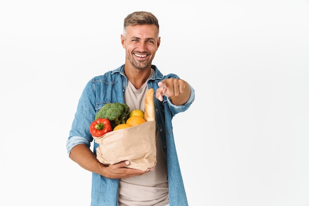 unrasierter Kerl in Jeanshemd zeigt mit dem Finger, während er einen Lebensmittelbeutel isoliert auf weiß hält