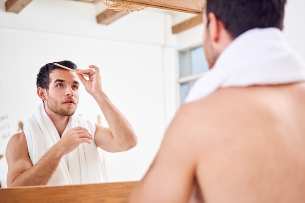 Unrasierter Mann, der sich die Haare kämmt, während er mit weißem Handtuch am Hals in der Nähe des Spiegels im Badezimmer steht