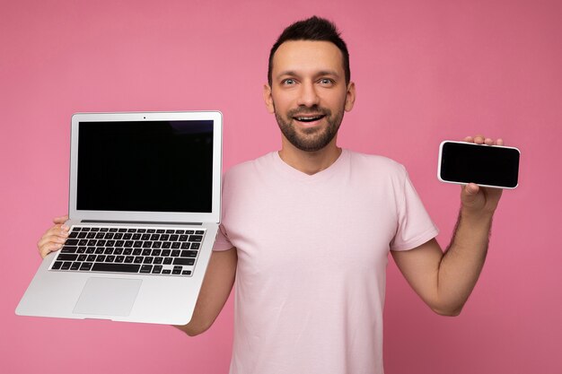 Unrasierter Mann des schönen Brunet, der Laptop-Computer und Handy hält, die Kamera im T-Shirt auf lokalisiertem rosa Hintergrund betrachten.