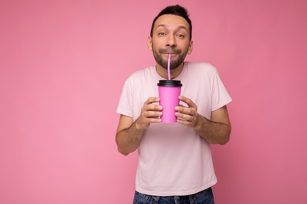 unrasierter Mann mit Bart, der weißes T-Shirt trägt, lokalisiert über rosa Hintergrundwand, die Papierkaffeetasse trinkt und Spaß hat.
