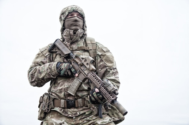 Unregelmäßiger Militärsöldner, Kommando-Saboteur in Tarnjacke, Kapuze auf dem Kopf tragen, bewaffnetes Dienstgewehr, taktischen Rucksack während des Marsches tragend, auf Distanz schauend