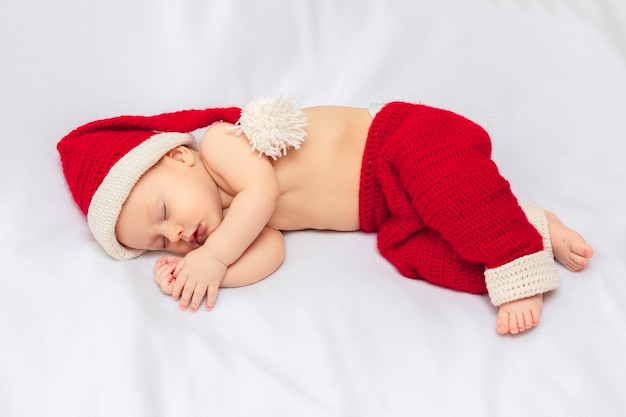 Foto unschuldiges baby, das handgemachten gehäkelten roten weihnachtselfenhut und eine hose trägt und auf weißer fleecedecke schläft