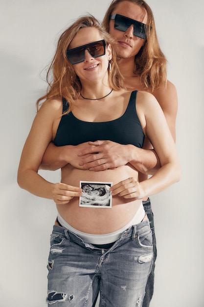 Unsere Familie Nahaufnahme eines jungen Mannes, der seine schöne schwangere Frau umarmt und ihren Bauch berührt, während sie ein Sonogrammfoto ihres Kindes hält