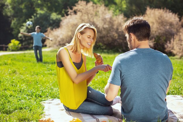 Unsere Freizeit. Fröhliche blonde Frau, die mit ihrem Ehemann und ihrem Kind spricht, die im Hintergrund spielen