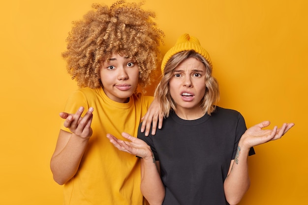 Foto unsichere frauen, die handflächen ausbreiten, stehen empört, können keine entscheidung treffen, zucken die schultern, tragen lässige t-shirts, die über gelbem hintergrund isoliert sind. unsichere, zögernde weibliche models fühlen sich in frage gestellt und ahnungslos