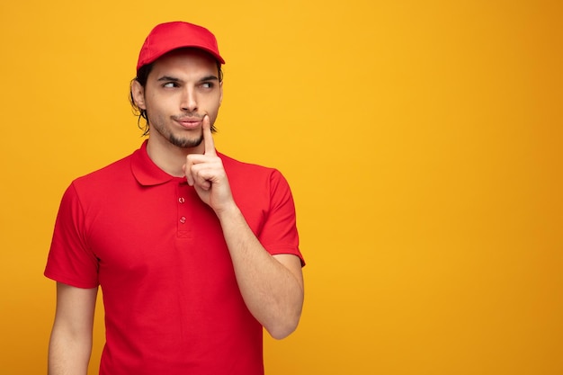 Unsicherer junger lieferbote, der uniform und kappe trägt, die das gesicht berühren, das die seite lokalisiert auf gelbem hintergrund mit kopienraum betrachtet