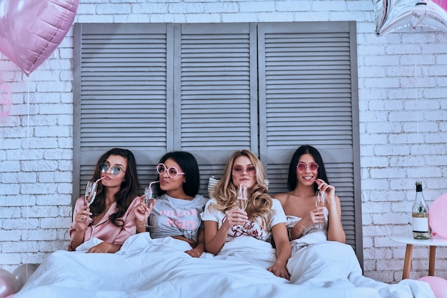 Foto unter freunden einfach du selbst sein! vier attraktive junge frauen im pyjama trinken cocktails, während sie im bett liegen