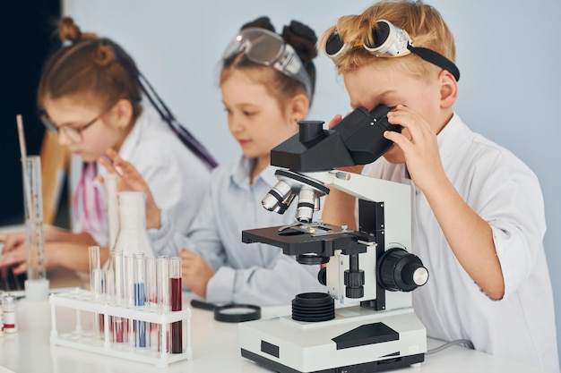 Unter Verwendung des Mikroskops Kinder in weißen Kitteln spielen einen Wissenschaftler im Labor, indem sie Ausrüstung verwenden